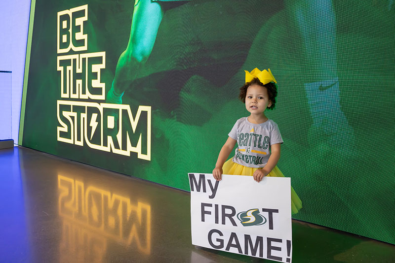 Toddler holding sign that reads "My First Game" in front of Seattle Storm "Be The Storm" Activation at Climate Pledge Arena