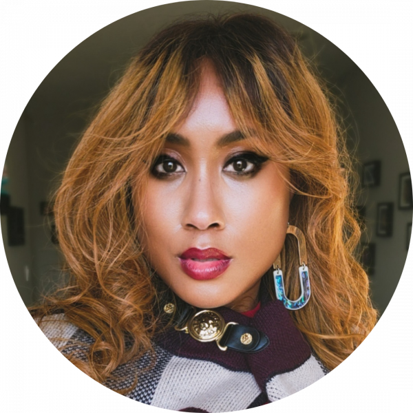 Photo of Lanaé Evans with blonde curled hair, wearing black eyeliner, burgundy lipstick, abalone and gold earrings, and a maroon, navy blue and cream cape with a black leather and gold clasp