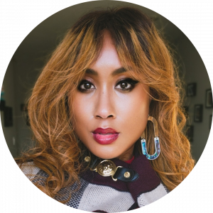 Photo of Lanaé Evans with blonde curled hair, wearing black eyeliner, burgundy lipstick, abalone and gold earrings, and a maroon, navy blue and cream cape with a black leather and gold clasp